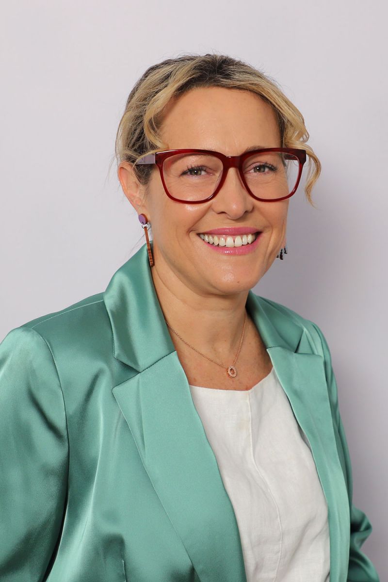 Portrait of smiling blonde middle-aged white female, wearing red glasses, green silk jacket and white blouse.